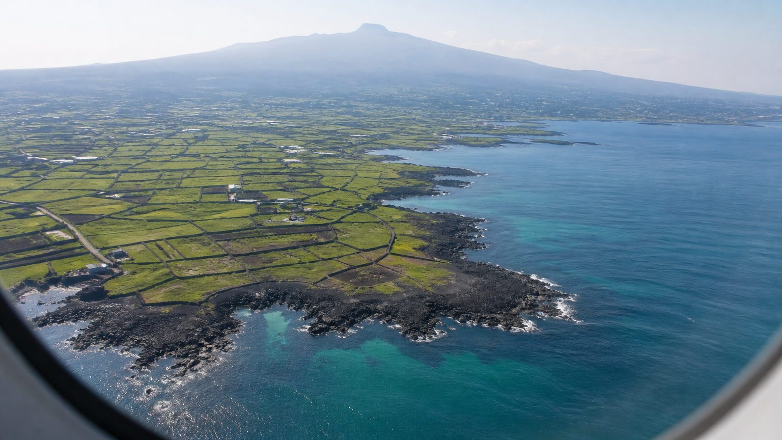 Jeju on approach — basalt coast, stone-walled fields, Hallasan in the haze.